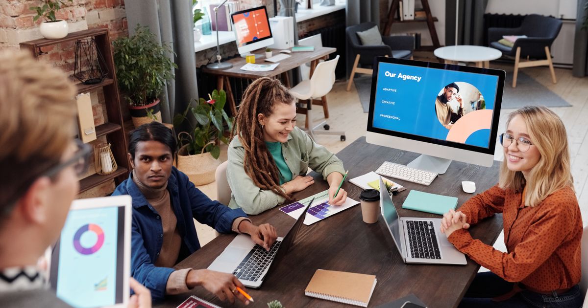 Equipe de agência reunida em mesa de trabalho com laptops e tela de apresentação