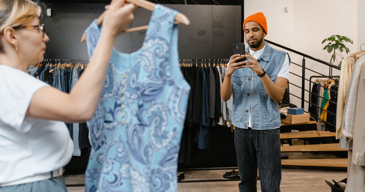 Numa loja de roupas, um rapaz fotografa com o celular o vestido que uma colega segura