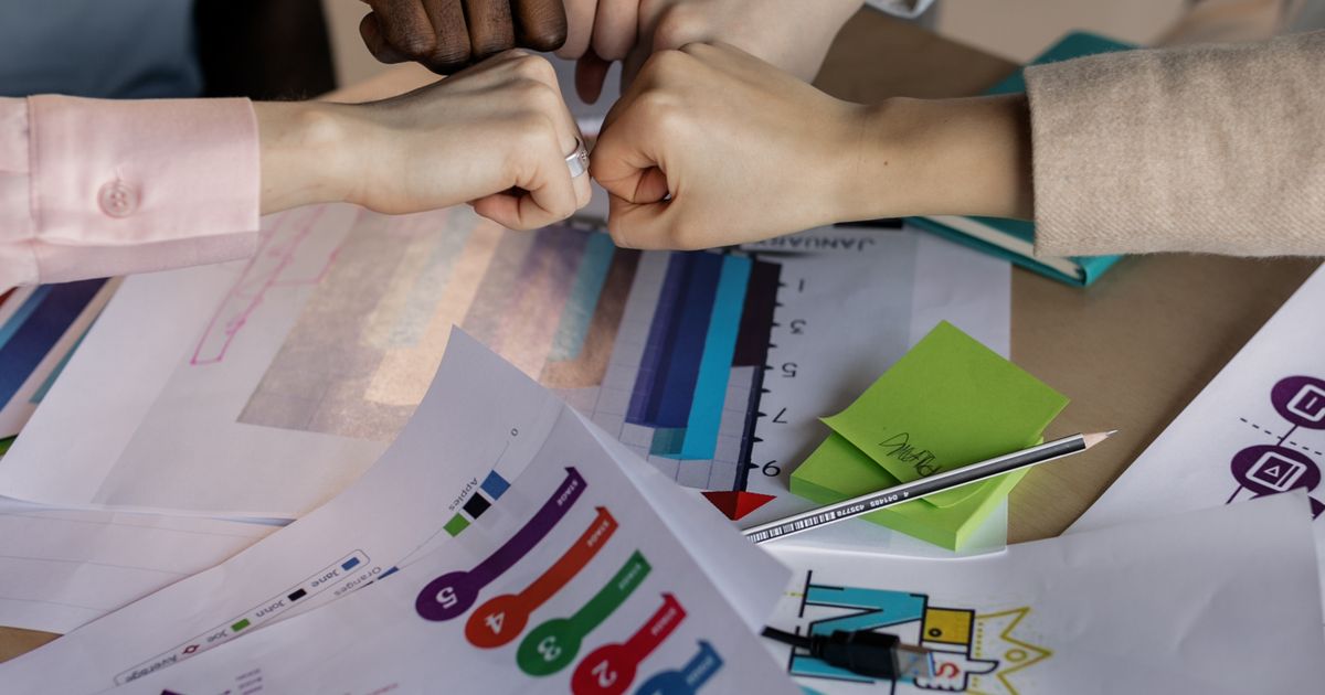 Mãos de uma equipe se cumprimentando sobre uma mesa coberta de gráficos, rankings e anotações